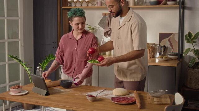 Girl with prosthetic arm viewing pizza recipe on digital tablet, boyfriend putting plate with tomatoes and fresh greens on table and joining her, he holding vegetable choosing cutting style