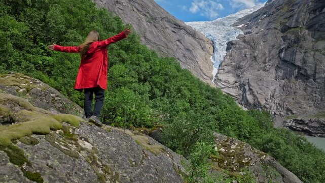 Female tourist standing with arms outstretched near Briksdalsbreen in Norway. Scenic glacier, cliffs and forest surround her during an inspiring summer travel moment.