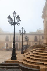 Vintage street lamps illuminate stone steps in a foggy square in Enna, Sicily, showcasing historic architecture and a serene atmosphere in the early morning light