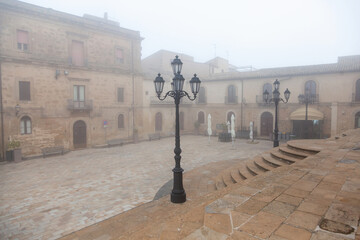 Foggy square in Enna, Sicily, featuring historic stone buildings, a decorative lamppost, and cobblestone pavement, creating a serene atmosphere in the early morning light