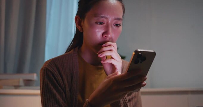 Stressed Asian woman looking at smartphone with worried and tearful expression at night.