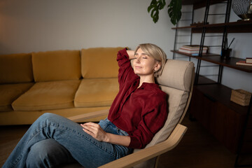 Serene middle aged woman relaxing at home, comfortably resting in soft chair in cozy living room enjoying calm moments. Peaceful female taking time to rest, sits with closed eyes and dreaming