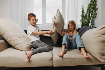 Two happy children having a pillow fight on a beige sofa in a bright living room with natural light...