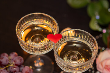 Two champagne glasses with red fabric heart and grapes for Valentine's Day on dark background with candles