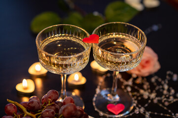 Two champagne glasses with red fabric heart and grapes for Valentine's Day on dark background with candles