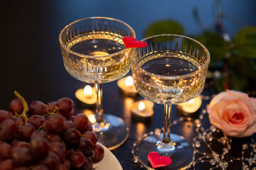 Two champagne glasses with red fabric heart and grapes for Valentine's Day on dark background with candles