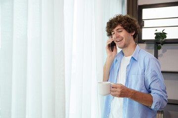 Fototapeta premium young man holding a cup of coffee and talking on smartphone beside window