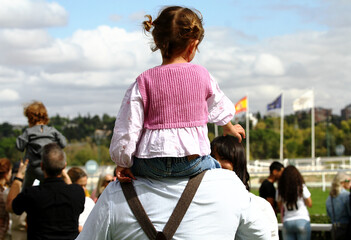 Ni&ntilde;a subida a hombros arriba de su padre a caballito con p&uacute;blico y espectador en evento al aire libre