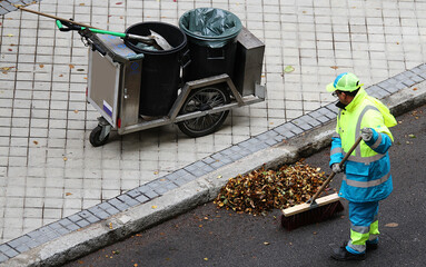 Barrendero municipal con escoba y cubo barriendo hojas del suelo en la calle con carro y recogedor
