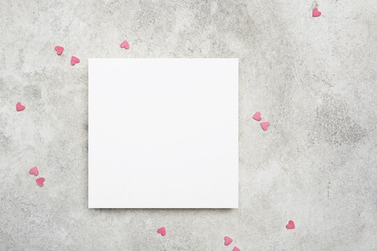 Blank square greeting or invitation paper card mockup with pink envelope and small hearts on grey stony background, copy space for card design