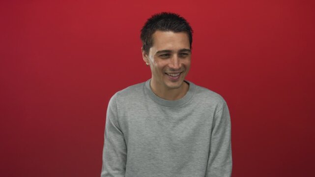 Young hispanic man smiling confidently against a vibrant red background wearing a casual grey sweater, conveying a relaxed and friendly demeanor.