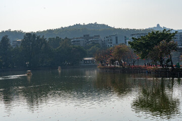 Obraz premium Calm lake with tree-lined shore, distant hills, and urban buildings under hazy sky - Huizhou West Lake