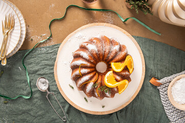 Top down view of a golden orange bundt cake decorated with citrus slices and fresh thyme sitting on a wooden serving board next to a green linen napkin and sugar sifter for a rustic kitchen scene