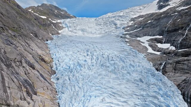 Wide drone view of Briksdalsbreen glacier descending through mountain valley in Norway. Blue ice contrasts with rocky slopes under bright summer conditions.