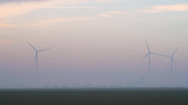 Distant wind farm partially obscured by thick fog at dawn, presenting a quiet rural environment where renewable energy infrastructure blends into the soft horizon