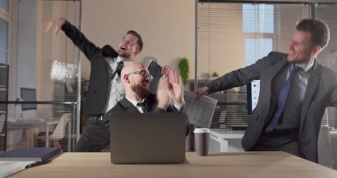 Businessmen team dance in office party celebration. Three men in suits groove with coworkers by a laptop, celebrating success and lifting workplace morale. Clear concept of teamwork and fun.
