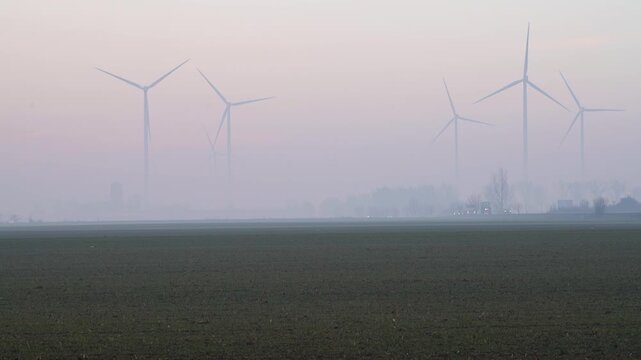 Wind turbines on the horizon across a calm rural plain, softened by fog and pastel sky tones, presenting a serene and sustainable energy landscape scene