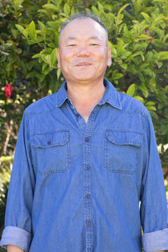 Senior Asian man standing in garden with green shrubs and red flowers, wearing blue denim shirt