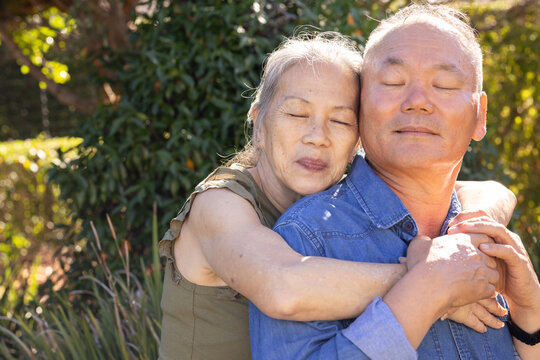 Senior Asian couple embracing, standing in sunlit garden with arms wrapped around shoulders