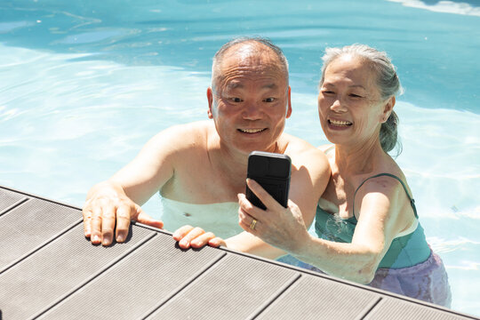 Senior Asian couple smiling and taking selfie in pool wearing swimsuits, holding phone and ring