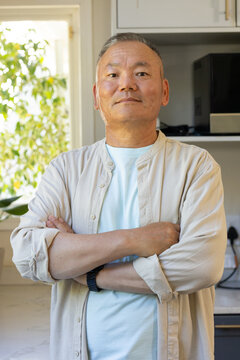 Senior Asian man standing arms crossed in kitchen by window plant wearing beige shirt and watch