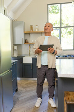 Senior Asian man standing in modern home kitchen holding tablet and checking wristwatch