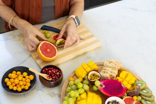Tropical fruit are being prepared on wooden cutting board on marble countertop, blue-handled knife