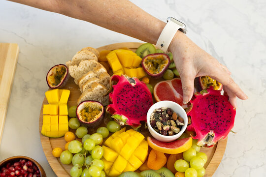 Wooden platter holding dragon fruit and mango on marble top, forearm with white smartwatch reaching