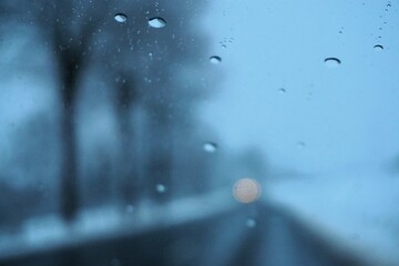 Abstraktes Motiv mit Wassertropfenmuster vor Schneelandschaft mit Orangegelbem Licht auf nasser Hauptstra&szlig;e zwischen Feldern und Baumreihe auf dem Land bei Eis, Frost und Schnee am Morgen im Winter