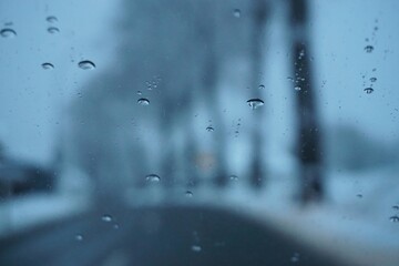 Abstraktes Motiv mit Wassertropfenmuster auf beschlagener Glasscheibe vor nasser Hauptstra&szlig;e zwischen Schneebedeckten Wiesen mit Reihe hoher B&auml;ume und Himmel auf dem Land Sturm und K&auml;lte  im Winter