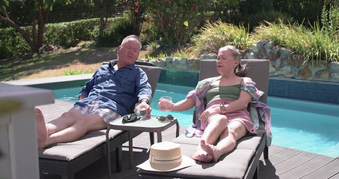 Asian couple lifting glasses in small toast, sipping, putting on sunglasses, relaxing on loungers