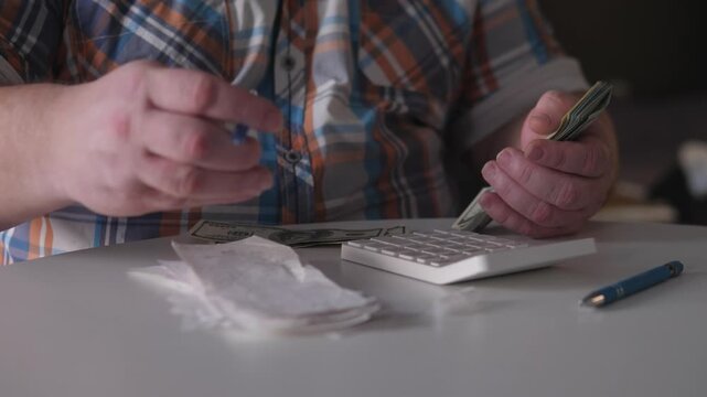 Worker Sorts Bills With Calculator, Man In Plaid Shirt Handles Cash Under Dim Indoor Lighting, Tired Man In Plaid Shirt Organizes Receipts And Bills After Work Shift Indoors