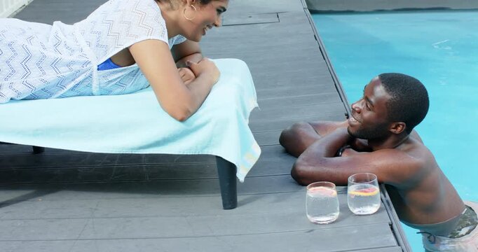 Eye contact sparking Diverse couple exchanging smiles on towel chair and pool deck near two glasses