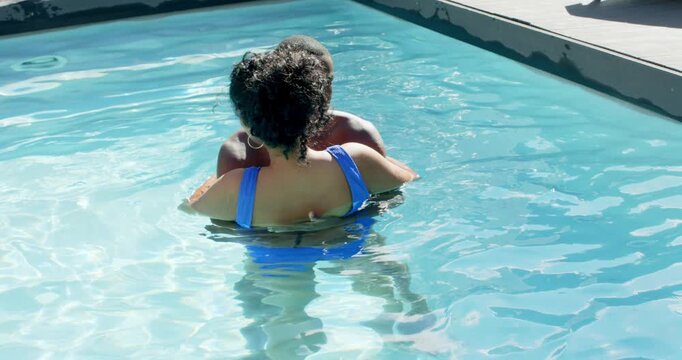 Diverse couple holding in pool, man adjusting hold, woman turning smiling, bonding in blue swimsuit