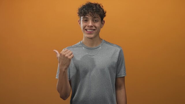 Man pointing thumb to side in studio wearing gray shirt and silver chain, smiling with relaxed pose; cheerful greeting.