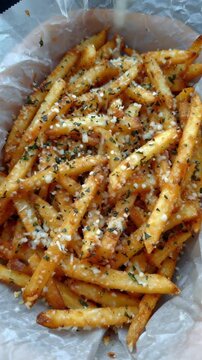 Close up top view of crispy french fries being sprinkled with parmesan cheese and herbs in paper lined tray
