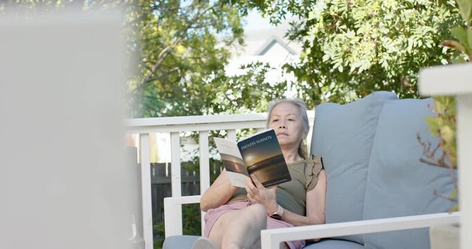 Senior Asian woman settling into cushion and reading paperback on deck sofa, relaxing at sunset
