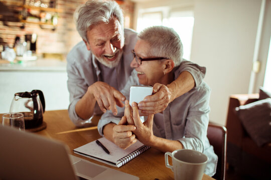 Senior couple laughing while using smartphone at home kitchen table