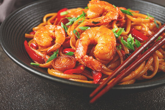 udon stir-fry shrimp noodles with red bell pepper and green onion, served in a black bowl, with chopsticks, dark background,