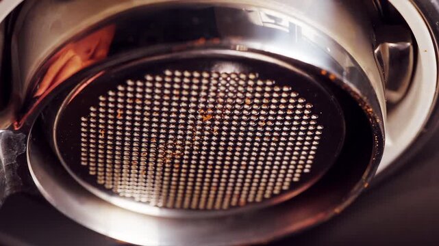 Close view of bottomless portafilter basket with coffee residue, macro perspective on metal filter surface during espresso setup, detailed professional brewing equipment scene