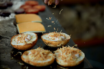 Burger buns topped with cheese and seasoning during food preparation, ideal for: catering services, restaurant operations, fast food production, commercial kitchen workflows.