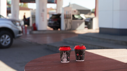 Coffee business at a gas station. Two paper cups of hot coffee on a street table against a blurred background of gas pumps and a car.