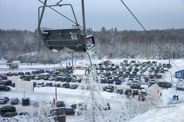 Panoramic aerial view of a car park at a ski resort surrounded by snow-covered forest. Two-seater chairlifts in the air. Winter morning.