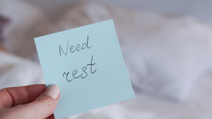 Hand holding sticky note with text "Need rest" on blurred bed background