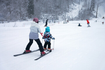 Technique for teaching young children to stand on alpine skis. A female skier holds a small child by a safety strap. They ski down the slope.