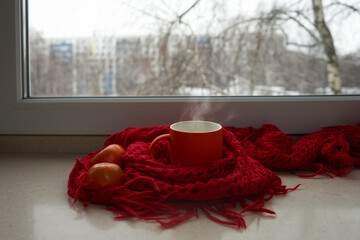 An orange mug with a hot drink, wrapped in a knitted red scarf, on the table by the window. Two tangerines nearby. Outside the window, a cold winter cityscape.
