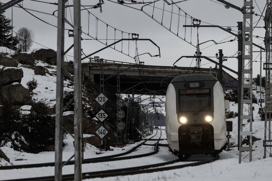 A passenger train speeds along snow-covered railway tracks through the countryside near the historic city of Avila, Spain, during a freezing winter day.