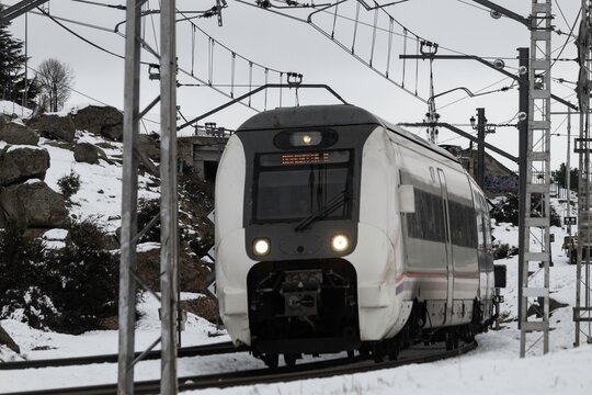 A passenger train speeds along snow-covered railway tracks through the countryside near the historic city of Avila, Spain, during a freezing winter day.