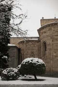 Heavy snow blankets the garden bushes outside the rear Romanesque exterior of San Pedro Church in Avila, Spain, highlighting the ancient stone architecture on a freezing winter day.