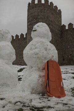 A lively winter scene at the northern stone fortifications of Avila, Spain, where locals and visitors play in the deep snow and build snowmen against the backdrop of the historic medieval walls.